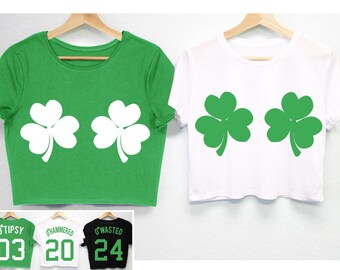 Peut inclure: Deux crop tops, un vert et un blanc, chacun avec un grand motif de trèfle. Le haut vert a deux trèfles blancs, tandis que le haut blanc a deux trèfles verts. En dessous, trois autres chemises avec du texte et des chiffres.