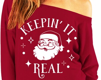 May include: Red off-the-shoulder sweatshirt with a white graphic of Santa Claus and the text "Keepin' it real."