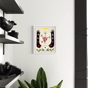 May include: A framed illustration of a stylized heart pierced by swords, with blood dripping from the wounds. The heart is surrounded by two black arms with red stars, and a crescent moon and eye are above the heart. The background is a light beige color.