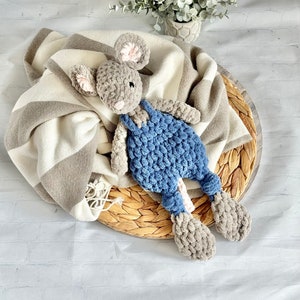 May include: A handmade crochet mouse toy with gray fur, pink inner ears, and blue overalls. The plush toy is lying on a striped blanket and a woven tray, with a small potted plant in the background. The toy is soft and cuddly.