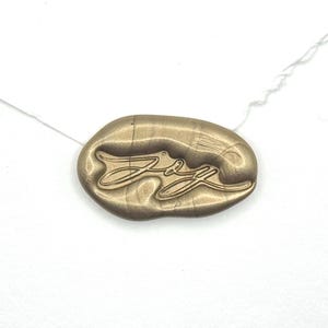 May include: An oval-shaped metallic gold wax seal featuring the word Joy in a cursive script font. The seal has a smooth, slightly irregular edge and a raised surface texture, centered on a plain white background with a thin white thread visible.