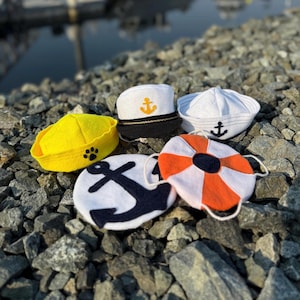May include: A collection of five felt nautical-themed accessories including a yellow hat with a black paw print, a white captain's hat with a gold anchor, a white sailor hat with a black anchor, a white disc with a black anchor, and a life ring.
