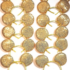 Puede incluir: Un conjunto de doce diademas doradas, cada una con dos orejas circulares cubiertas de lentejuelas doradas brillantes. Las diademas están dispuestas en dos columnas verticales de seis, mostrando la textura metálica y el brillo de las lentejuelas.