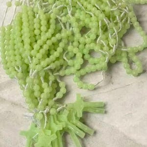 May include: A collection of light green rosary beads with small, round beads strung on white string. Several cross-shaped pendants are also visible. The beads and crosses have a soft, glowing appearance.