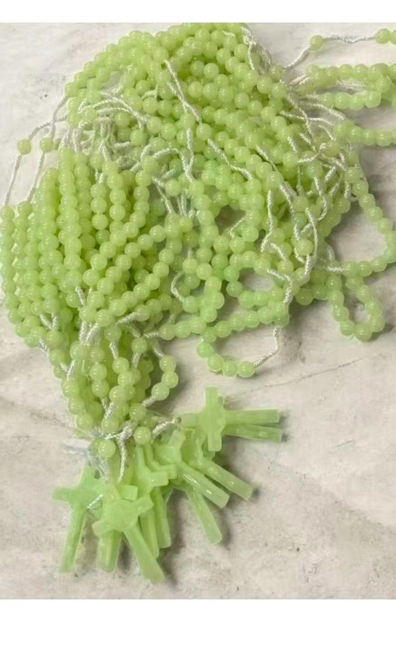 May include: A collection of light green rosary beads with small, round beads strung on white string. Several cross-shaped pendants are also visible. The beads and crosses have a soft, glowing appearance.