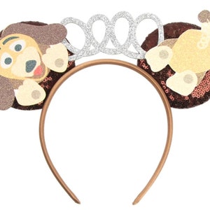 May include: Brown headband with a sparkly brown and beige dog ear on each side. The headband has a silver crown on top.