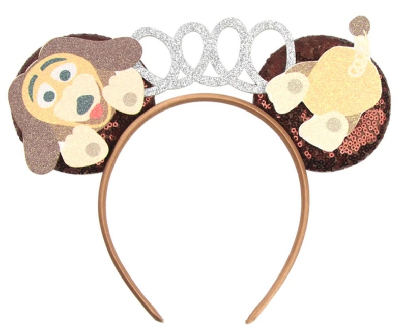 May include: Brown headband with a sparkly brown and beige dog ear on each side. The headband has a silver crown on top.