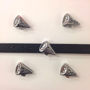 Peut inclure: Cinq pendentifs de charme argentés avec des strass et le mot "CHEER" sur chacun. Les breloques sont en forme de mégaphones.