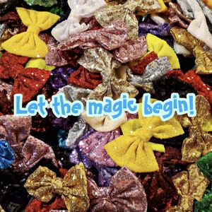 Può includere: Una collezione di fiocchi per capelli scintillanti in vari colori, tra cui oro, argento, rosso, blu e verde. I fiocchi sono fatti di paillettes e sono disposti in una pila. Il testo "Let the magic begin!" è scritto in lettere blu su uno sfondo bianco.