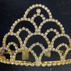 May include: A gold crown with a decorative design of clear rhinestones. The crown has a rounded top and a flat base.