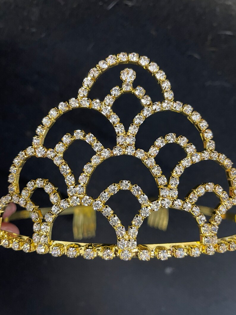 May include: A gold crown with a decorative design of clear rhinestones. The crown has a rounded top and a flat base.