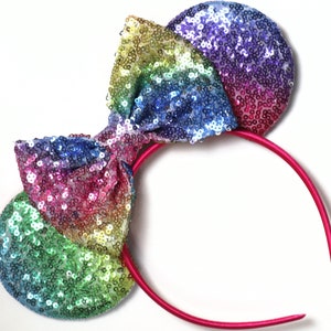 May include: Rainbow sequined headband with a large bow. The headband is pink and the bow is made of iridescent sequins in shades of green, blue, purple, pink, and yellow.