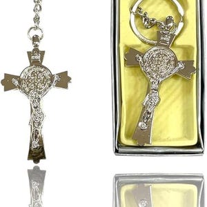 May include: Silver-colored crucifix keychain with a detailed depiction of Jesus on the cross. The keychain is displayed next to a boxed version, both featuring a religious theme. The cross has intricate detailing and a shiny finish.
