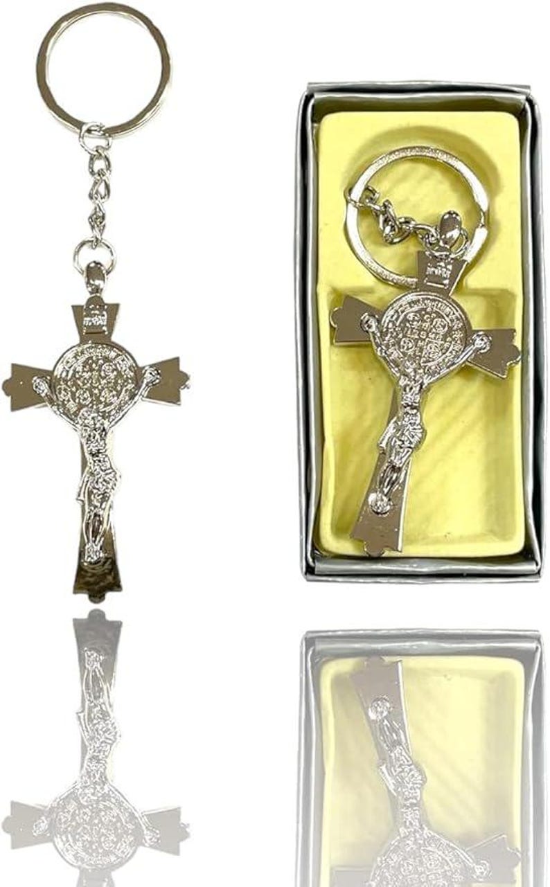 May include: Silver-colored crucifix keychain with a detailed depiction of Jesus on the cross. The keychain is displayed next to a boxed version, both featuring a religious theme. The cross has intricate detailing and a shiny finish.