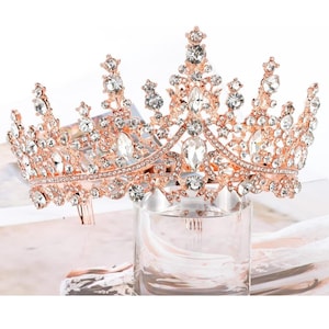 Pode incluir: Coroa em tom de ouro rosa com cristais transparentes e brilhantes. A tiara apresenta um design detalhado com cristais de v&aacute;rios tamanhos, dispostos em um padr&atilde;o floral e geom&eacute;trico. A coroa &eacute; exibida em um cilindro de vidro transparente.