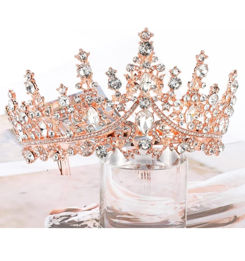 Pode incluir: Coroa em tom de ouro rosa com cristais transparentes e brilhantes. A tiara apresenta um design detalhado com cristais de v&aacute;rios tamanhos, dispostos em um padr&atilde;o floral e geom&eacute;trico. A coroa &eacute; exibida em um cilindro de vidro transparente.