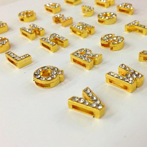 May include: A set of gold-colored letter charms with clear rhinestones. The letters are all uppercase and are arranged in a random order.