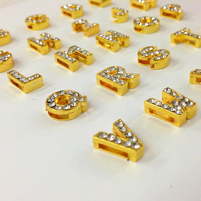 May include: A set of gold-colored letter charms with clear rhinestones. The letters are all uppercase and are arranged in a random order.