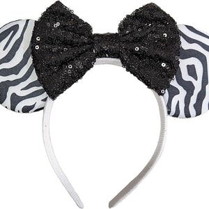 May include: A headband featuring two circular ears with a black and white zebra stripe pattern. Centred between the ears is a large, prominent bow covered in black sequins that catch the light. The headband frame itself is a plain white colour.