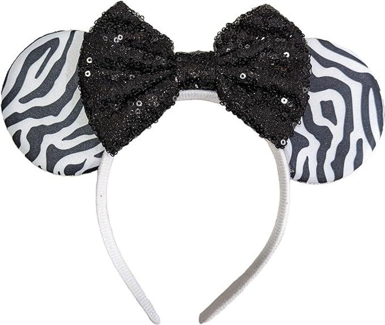 May include: A headband featuring two circular ears with a black and white zebra stripe pattern. Centred between the ears is a large, prominent bow covered in black sequins that catch the light. The headband frame itself is a plain white colour.