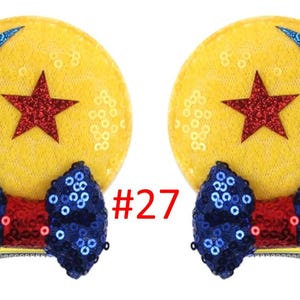 Puede incluir: Dos lazos para el pelo amarillos con lentejuelas con un dise&ntilde;o de estrella roja y media luna azul. Cada lazo tiene un lazo azul con lentejuelas y un centro rojo. El n&uacute;mero "27" est&aacute; impreso en rojo.
