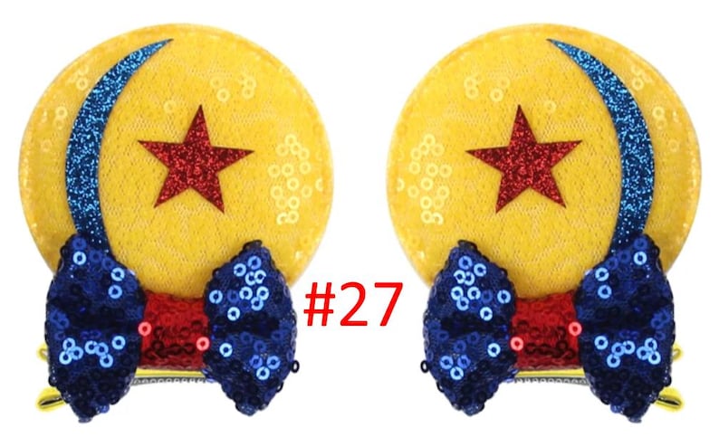 Puede incluir: Dos lazos para el pelo amarillos con lentejuelas con un dise&ntilde;o de estrella roja y media luna azul. Cada lazo tiene un lazo azul con lentejuelas y un centro rojo. El n&uacute;mero "27" est&aacute; impreso en rojo.