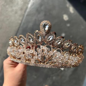 Pode incluir: Uma tiara dourada com cristais e pedras preciosas transparentes e brilhantes. A coroa apresenta uma gema central em forma de gota e pedras ovais e redondas menores. O design inclui intrincados padr&otilde;es florais e de folhas.