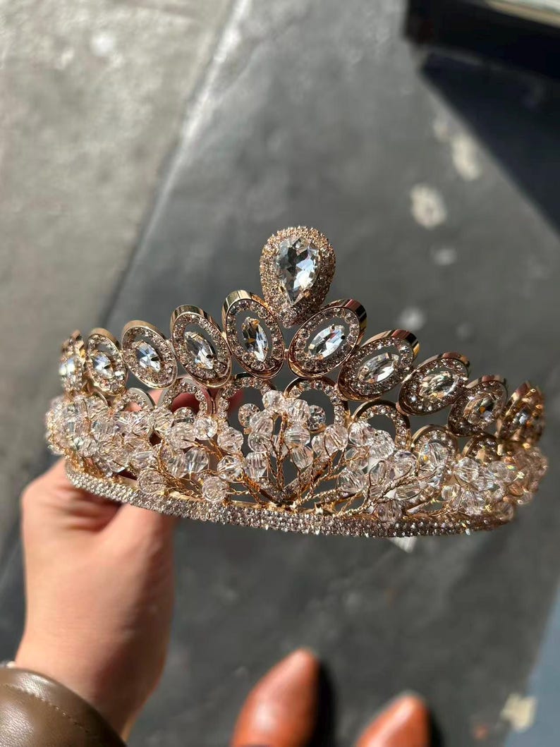 Pode incluir: Uma tiara dourada com cristais e pedras preciosas transparentes e brilhantes. A coroa apresenta uma gema central em forma de gota e pedras ovais e redondas menores. O design inclui intrincados padr&otilde;es florais e de folhas.