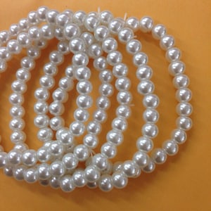 May include: A continuous strand of round, white, lustrous beads arranged in a circular pattern. The beads have a smooth, reflective surface and are strung together on a thin, white cord, creating a uniform, circular loop of white spherical beads.