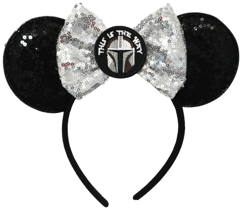 May include: Black and silver sequined Minnie Mouse ears headband with a silver bow and a circular patch featuring a Mandalorian helmet with the text "This is the way".