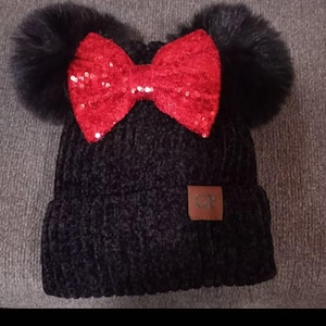 Puede incluir: Gorro de punto negro con un lazo rojo de lentejuelas y dos pompones de piel sintética negros. Una etiqueta de cuero marrón con un logotipo está cosida en el lateral. El gorro está hecho de un material suave y texturizado.