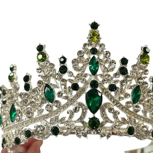 Pode incluir: Uma coroa prateada com detalhes em cristal verde esmeralda e transparente. A coroa apresenta um design sim&eacute;trico com grandes pedras verdes em forma de gota e cristais redondos menores. O estilo &eacute; ornamentado e elegante.