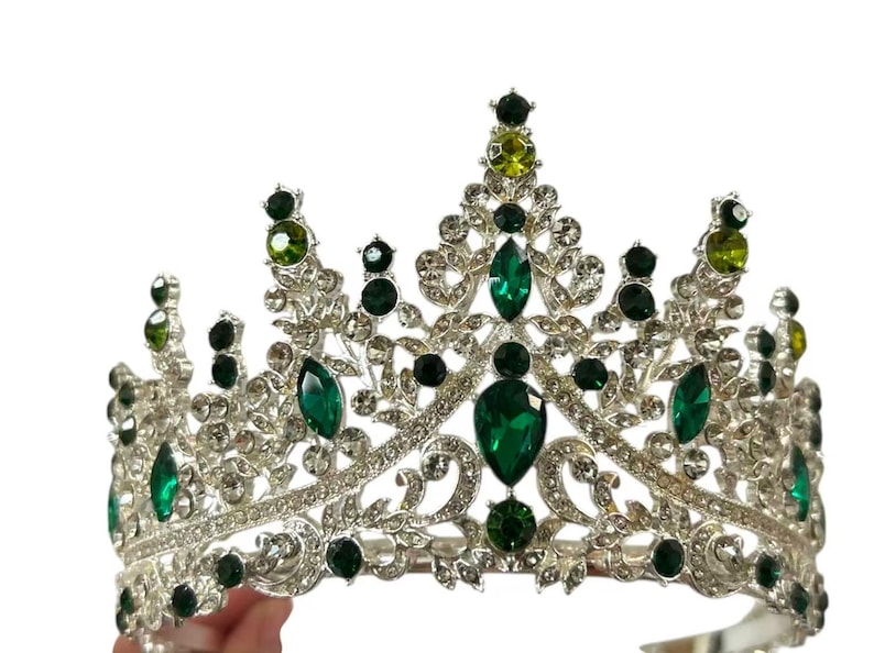 Pode incluir: Uma coroa prateada com detalhes em cristal verde esmeralda e transparente. A coroa apresenta um design sim&eacute;trico com grandes pedras verdes em forma de gota e cristais redondos menores. O estilo &eacute; ornamentado e elegante.