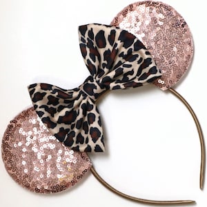 May include: A headband featuring two circular ears covered in shimmering rose gold sequins. Centered between the ears is a large, fabric bow with a tan, brown, and black leopard print pattern. The headband frame is a thin, metallic gold band.