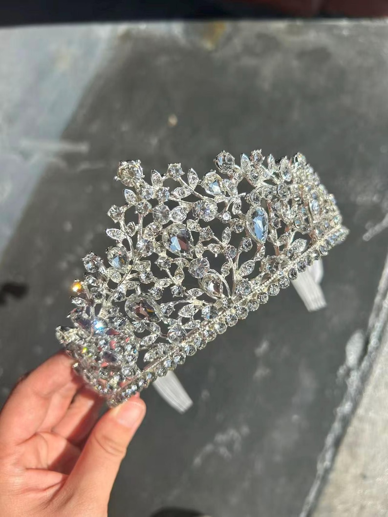 Pode incluir: Uma tiara prateada com um design floral, adornada com numerosos cristais claros e brilhantes. A coroa apresenta um delicado padr&atilde;o vazado e &eacute; presa por faixas brancas. A tiara &eacute; projetada para eventos formais.