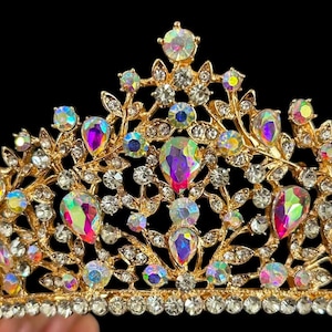 Pode incluir: Uma coroa dourada com pedras preciosas iridescentes em forma de gota e redondas. A coroa tem um design floral com strass redondos e transparentes. O fundo &eacute; preto.