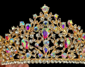 Puede incluir: Una corona dorada con piedras preciosas iridiscentes en forma de lágrima y redondas. La corona tiene un diseño floral con brillantes redondos y transparentes. El fondo es negro.