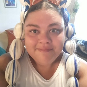 Pode incluir: Um par de orelhas com padr&atilde;o branco e azul numa bandolete com um la&ccedil;o central de lantejoulas cor de laranja. A bandolete &eacute; usada com duas extens&otilde;es de cabelo branco compridas, estilizadas em tran&ccedil;as de bolha com detalhes azuis.