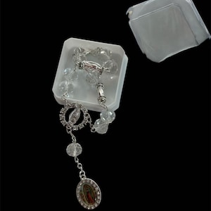 May include: A clear crystal rosary with silver-toned accents, displayed in a white plastic box. The rosary features a detailed pendant with a religious image and clear, faceted beads. The box lid is slightly ajar, revealing the contents.