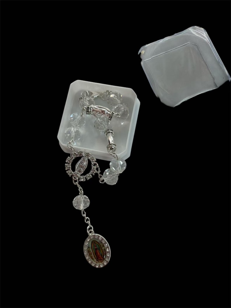 May include: A clear crystal rosary with silver-toned accents, displayed in a white plastic box. The rosary features a detailed pendant with a religious image and clear, faceted beads. The box lid is slightly ajar, revealing the contents.