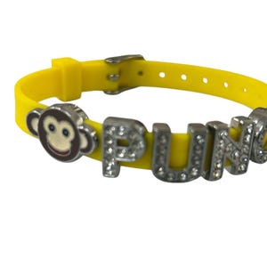 May include: Yellow rubber bracelet with a silver-colored buckle and the word "PUNKY" spelled out in silver letters encrusted with rhinestones. A monkey face charm is on the left side of the bracelet.