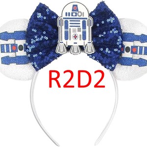 Pode incluir: Uma tiara com duas orelhas circulares brancas com gr&aacute;ficos de droide em azul e preto, um la&ccedil;o central de lantejoulas azul-escuro e um emblema do personagem R2D2. O texto R2D2 est&aacute; impresso em vermelho abaixo do la&ccedil;o na tiara.