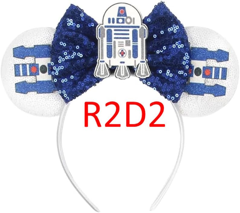 Pode incluir: Uma tiara com duas orelhas circulares brancas com gr&aacute;ficos de droide em azul e preto, um la&ccedil;o central de lantejoulas azul-escuro e um emblema do personagem R2D2. O texto R2D2 est&aacute; impresso em vermelho abaixo do la&ccedil;o na tiara.