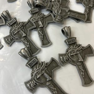 May include: A collection of silver-toned metal cross pendants. Each cross features a detailed depiction of a face with a crown of thorns and the inscription "INRI" at the top. The crosses are designed with a textured finish.
