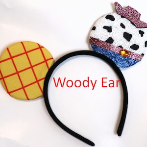 May include: A black headband with two felt ears. One ear is yellow with red squares and the other is white with black spots and a pink and purple cowboy hat. The headband says "Woody Ear".
