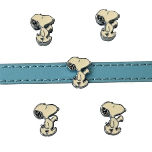 Puede incluir: Una correa de cuero azul claro con cinco dijes de Snoopy. Cada dije presenta una figura de Snoopy blanca con un contorno plateado. Los dijes están espaciados uniformemente a lo largo de la correa.