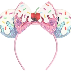 May include: A headband featuring two circular ears covered in light blue sequins with white icing and colourful sprinkles. A central pink sequined bow is topped with a white icing detail and a red glittery cherry with a green stem.