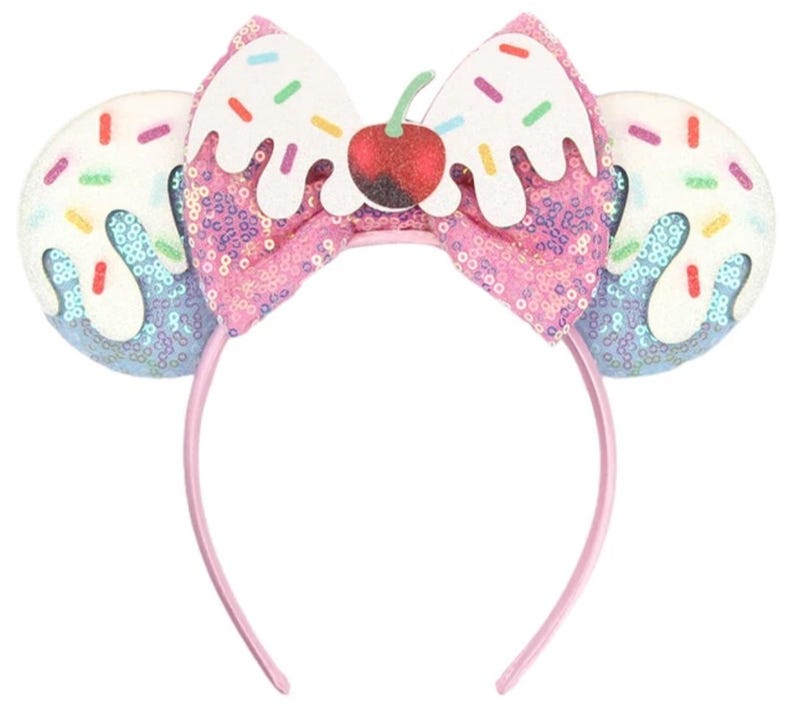 May include: A headband featuring two circular ears covered in light blue sequins with white icing and colourful sprinkles. A central pink sequined bow is topped with a white icing detail and a red glittery cherry with a green stem.