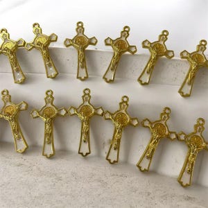 May include: A collection of twelve gold-toned metal crucifix pendants arranged in two rows. Each pendant features a white enamel inlay and a central circular medallion depicting a figure, with a detailed gold-toned figure of Jesus Christ.