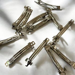 May include: A collection of silver-toned metal hair barrettes. The barrettes are rectangular with a curved design and a hinged clasp. They are scattered on a white surface.